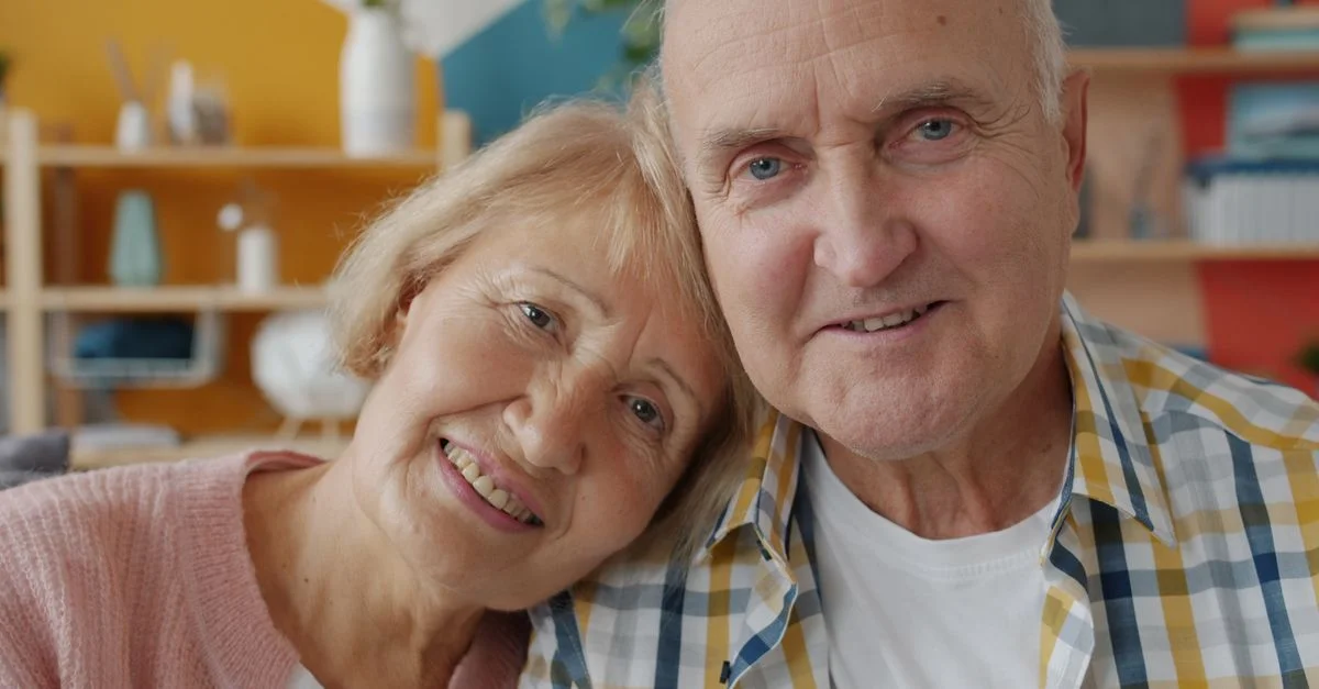 Un charmant couple de personnes âgées, souriant chaleureusement, est assis à l'intérieur et profite d'un agréable moment ensemble.