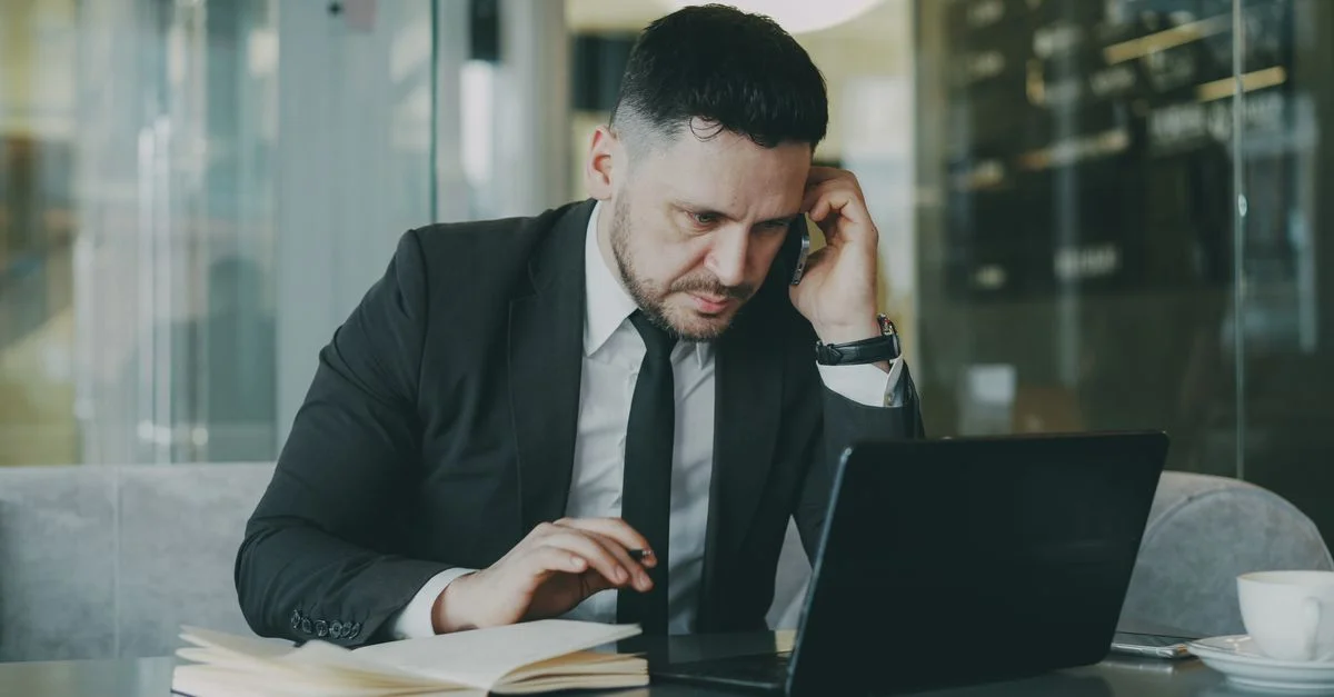 Homme d'affaires concentré en costume multitâche sur le téléphone et l'ordinateur portable dans un bureau moderne.