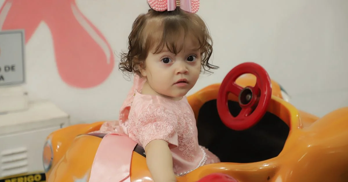 Une jolie petite fille portant une robe rose et un bandeau conduit une petite voiture orange à l'intérieur, montrant sa curiosité.